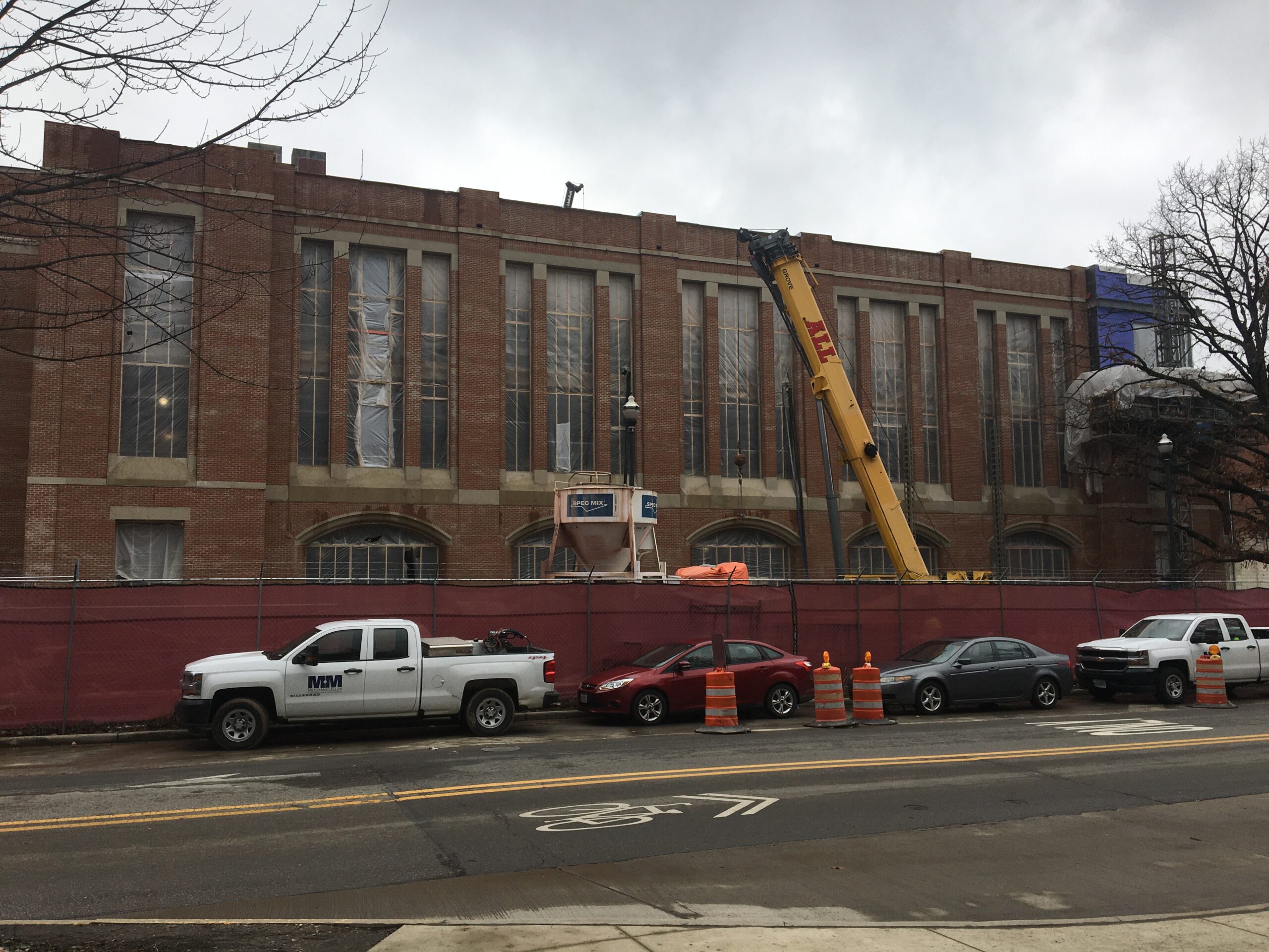 Large brick building with a crane in front of it, surrounded by vehicles on the road, traffic cones, a fence