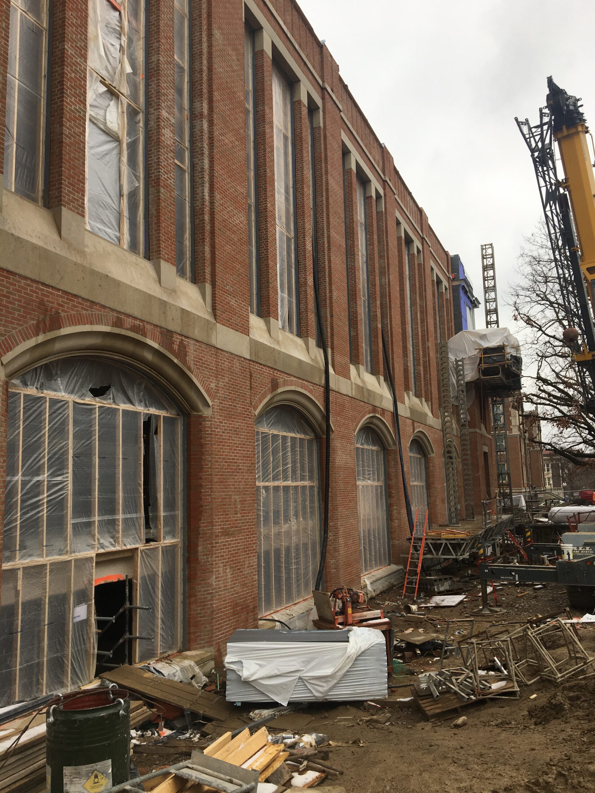 Large brick building with a crane in front of it, surrounded by trees, ladders, and other objects on the ground